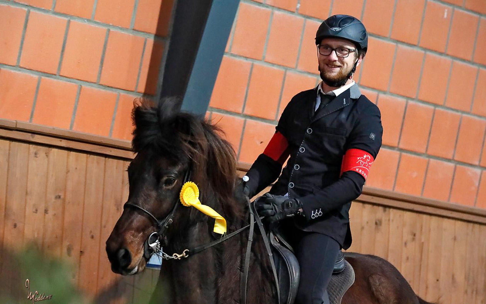 Fabian-Schrader-Labenslauf-0 Ein Reiter in Turnierkleidung sitzt auf einem dunkelbraunen Islandpferd mit einer gelben Schleife am Zaumzeug und lächelt.