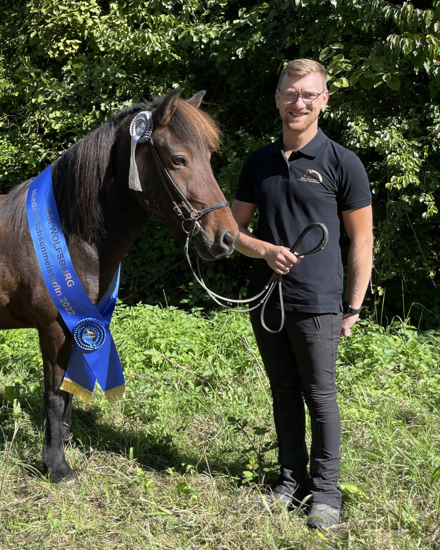 Ein Mann in schwarzem Poloshirt steht lächelnd neben einem prämierten Islandpferd mit blauer Siegerschärpe und silberner Schleife.