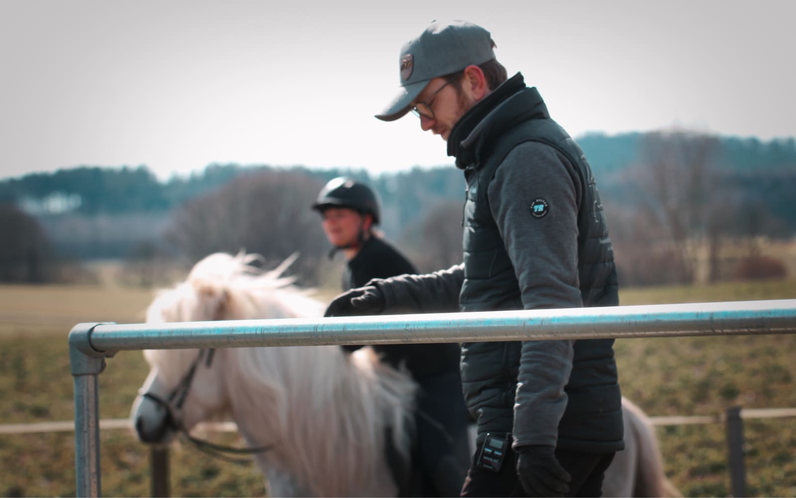 Ein Trainer in dunkler Kleidung und grauer Cap lehnt an einem Metallzaun, während im Hintergrund ein Reiter auf einem weißen Islandpferd unterwegs ist.